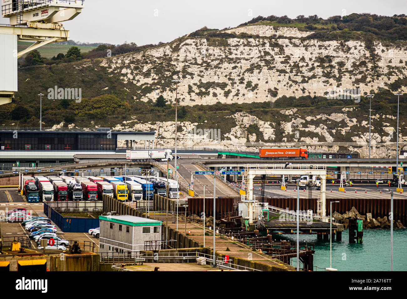 I camion si accalcano su più corsie per il controllo doganale al porto delle navi passeggeri di Dover, in Inghilterra, mentre un camion con lo slogan 'Sempre in orario' attraversa un ponte sopra di loro. Le scogliere bianche della città fanno da sfondo. Rimane poco spazio per manovrare nell'area ristretta del porto quando il traffico si ferma durante i controlli. Foto Stock