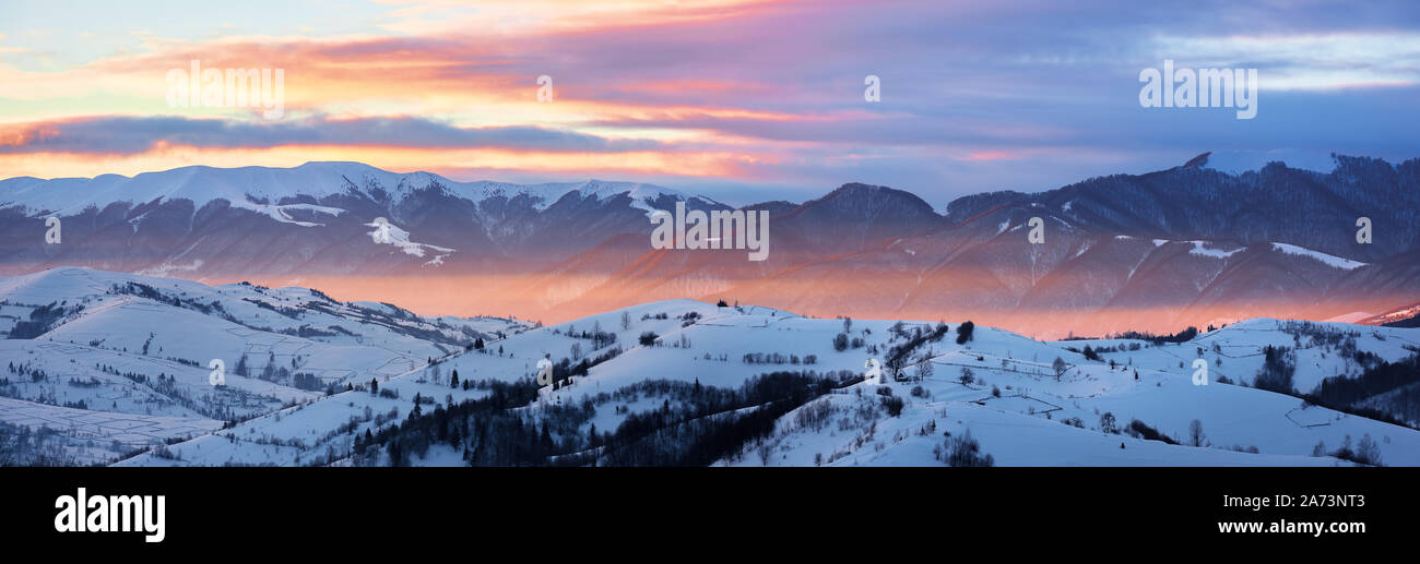 Stupendo panorama della campagna montuosa al sorgere del sole in inverno. coperta di neve area rurale su dolci colline glow in luce del sole di mattina. enorme montagna ri Foto Stock