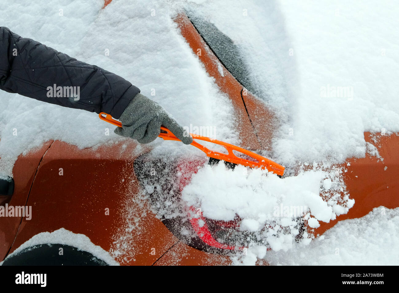 L'uomo cancella arancione auto da neve in una fredda giornata invernale dopo la nevicata. Spazzola in mano mans. Tanta neve su auto. Foto Stock