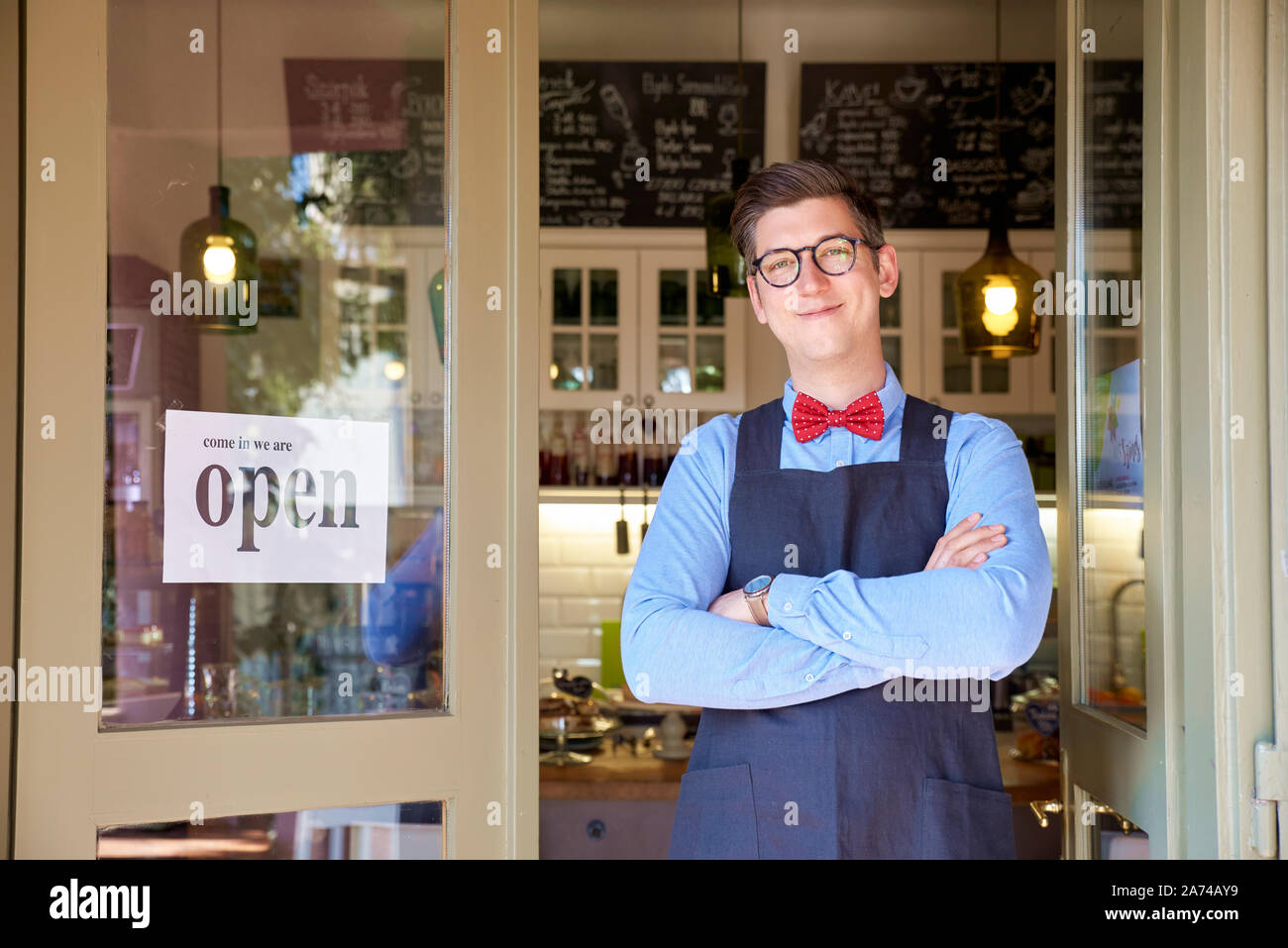 Ritratto di giovane titolare di una piccola azienda uomo in piedi con le braccia incrociate nella sua caffetteria ingresso. Foto Stock