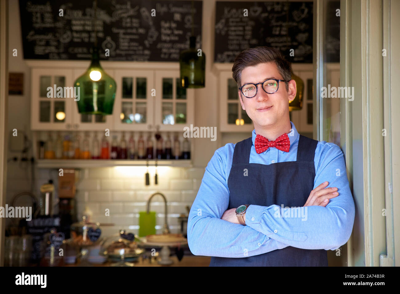 Ritratto di giovane titolare di una piccola azienda uomo in piedi con le braccia incrociate nella sua caffetteria ingresso. Foto Stock