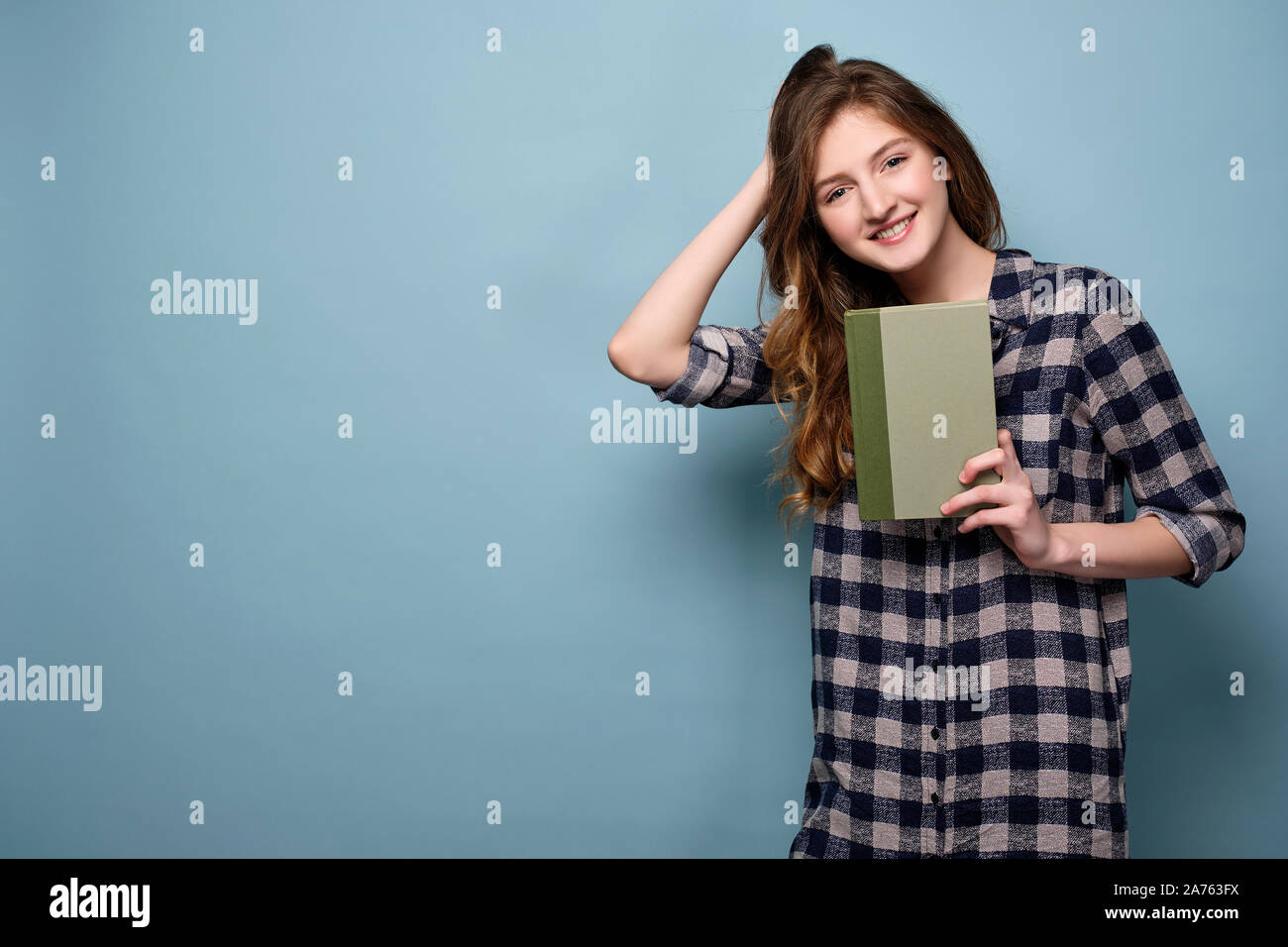 Una giovane ragazza in un abito di plaid sorge su uno sfondo blu e tiene un libro davanti a lui, esegue la sua mano attraverso i suoi capelli. Foto Stock