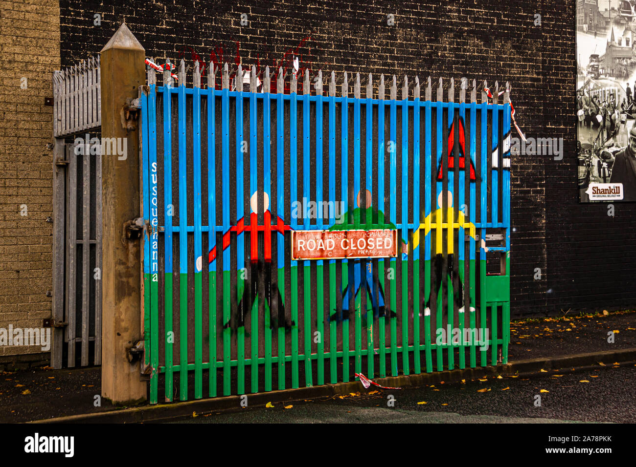 Un cancello dipinto di blu e verde sulla Muraglia Internazionale, Northumberland Street, Belfast, Irlanda del Nord, mostra tre figure stilizzate in rosso, verde e giallo che tengono insieme un cartello "Strada Chiusa"; la parola "Susquehanna" è dipinta verticalmente sul telaio del cancello. Foto Stock