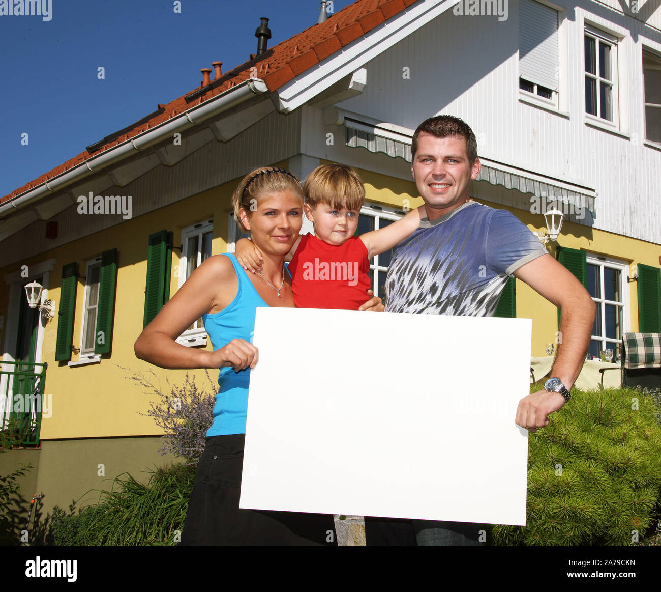 Junge Familie mit tipo neugekauftem vor Haus, hält weisses Schild, frei für beliebigen Testo, 30,35, 5 Jahre, signor:Sì Foto Stock