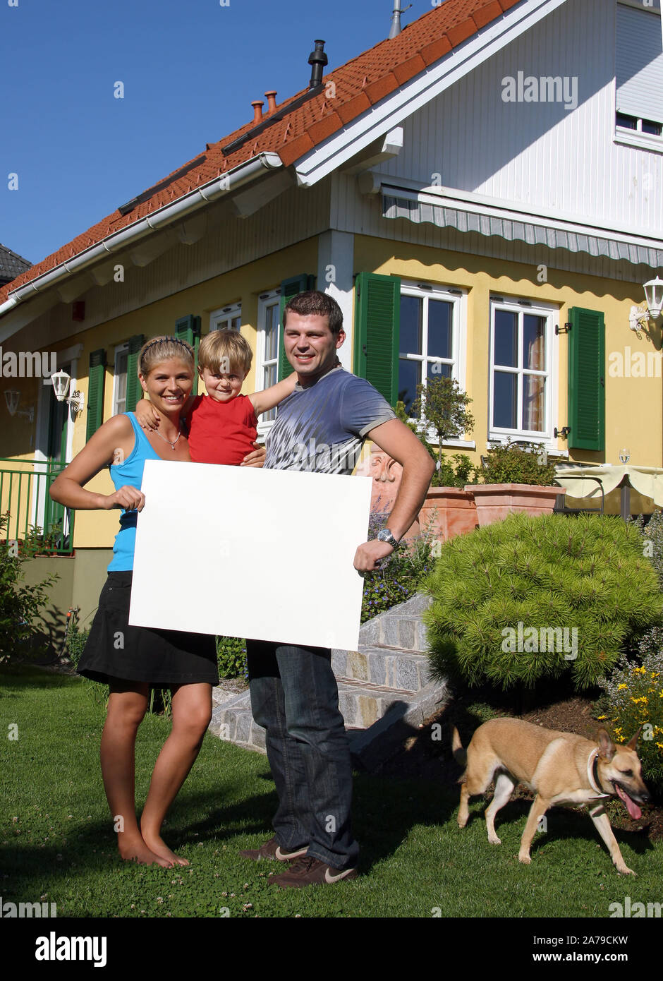 Junge Familie mit tipo neugekauftem vor Haus, hält weisses Schild, frei für beliebigen Testo, 30,35, 5 Jahre, signor:Sì Foto Stock