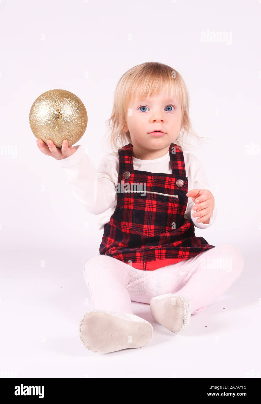 Piccola bionda toddler ragazza con grandi occhi blu in abito rosso gioca con la palla di Natale,seduta sul pavimento, sfondo bianco.ritratto, close-up isolato Foto Stock