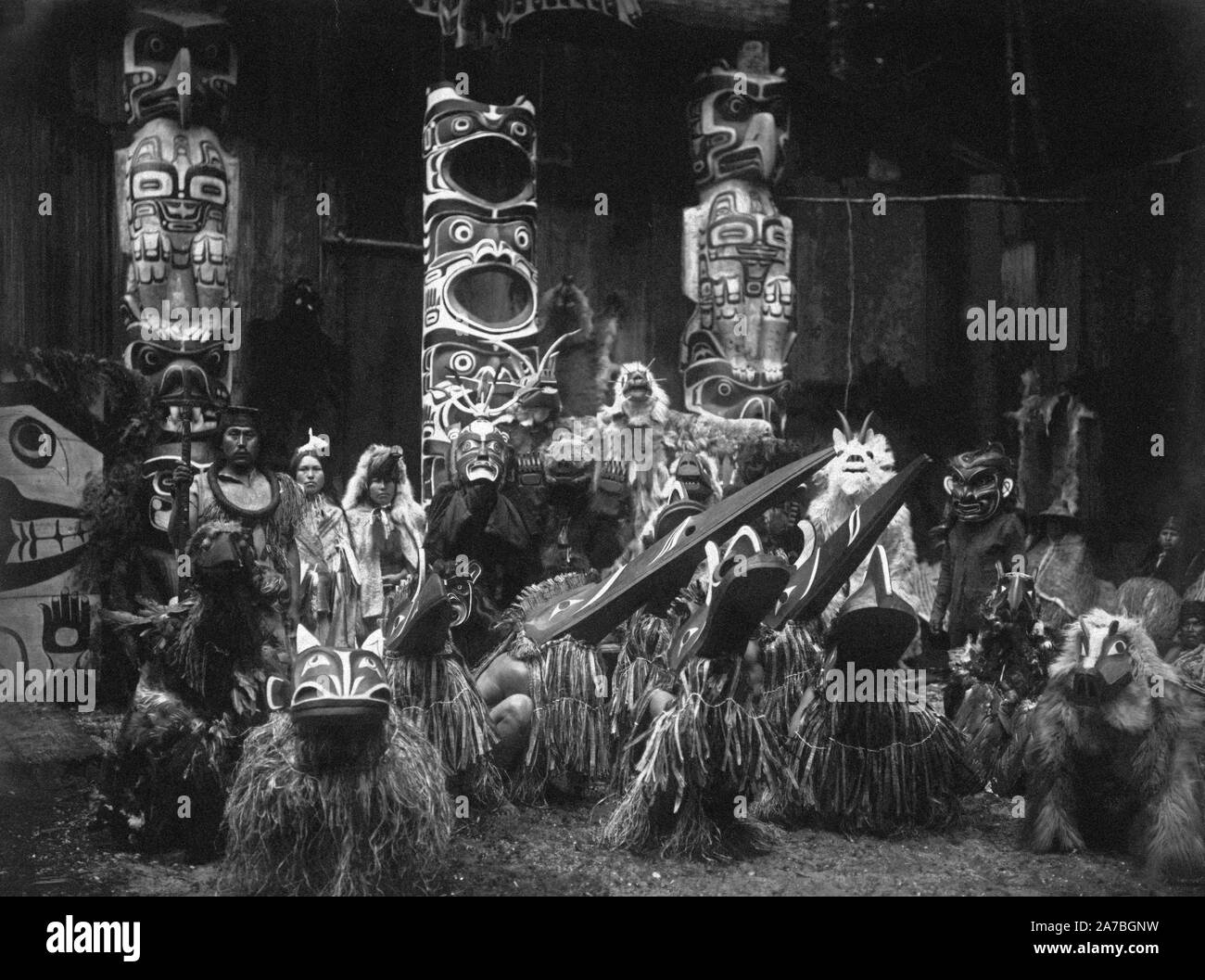 Durante la cerimonia d'inverno, il Kwakiutl ballerini indossano maschere e costumi, crouch in primo piano con gli altri dietro di loro. Il capo a sinistra contiene un altoparlante di personale. Tre totem in background. Foto Stock