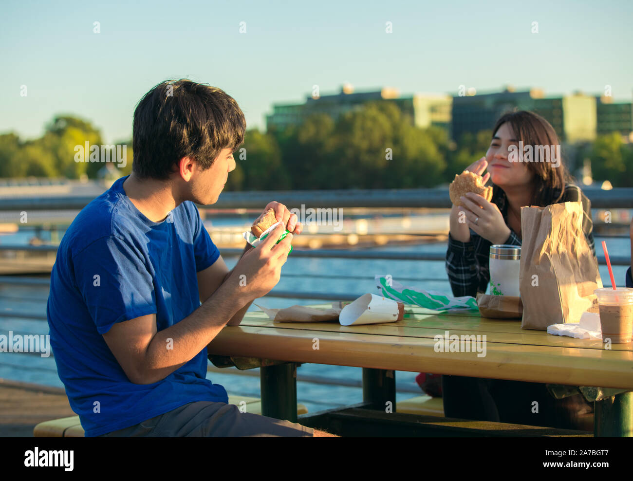 Due giovani asiatici biracial, caucasiche adulti seduti all'aperto sul lago di garda in area urbana di mangiare hamburger e parlare insieme al tavolo da picnic vicino al tramonto Foto Stock