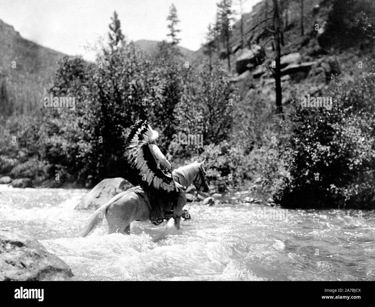 Edward S. Curtis nativi indiani americani - Bullchief, indossando warbonnet, attraversando i rapids poco profonde a cavallo ca. 1905 Foto Stock