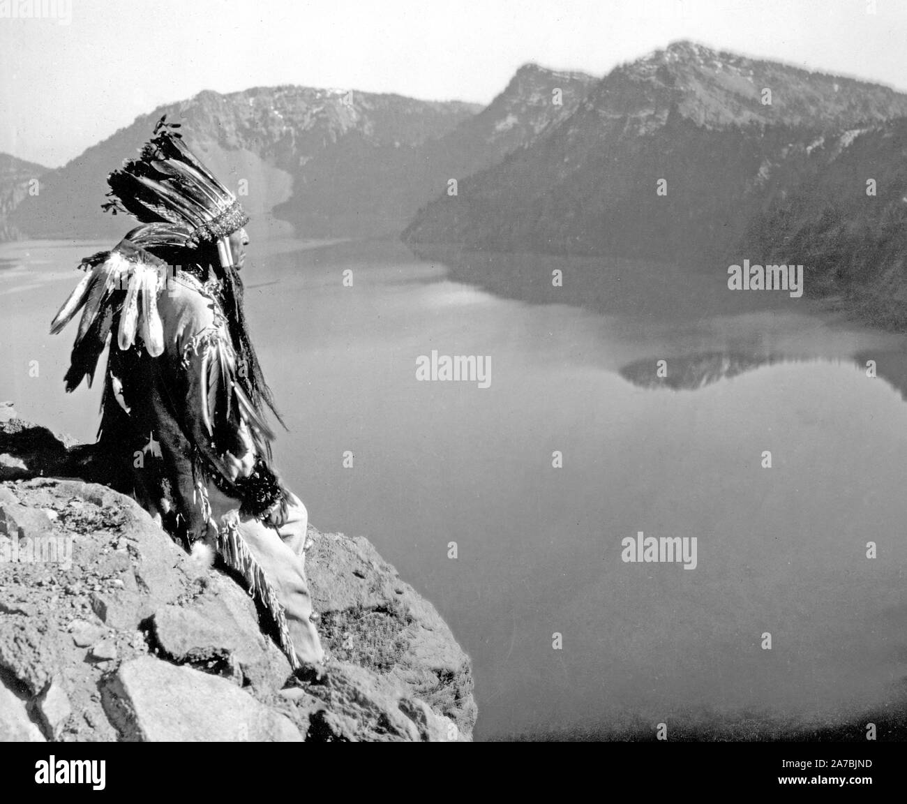 La fotografia mostra un American Indian uomo che indossa un headress, seduto su di una roccia overlocking di Crater Lake, Oregon Foto Stock