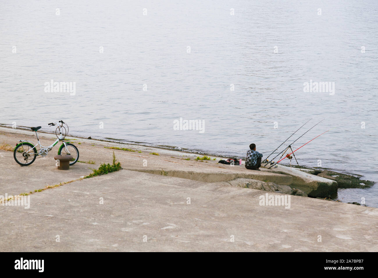 Un ricreative fisher uomo in attesa per la sua cattura da parte del fiume e la sua bicicletta vicino parcheggiata. Foto Stock