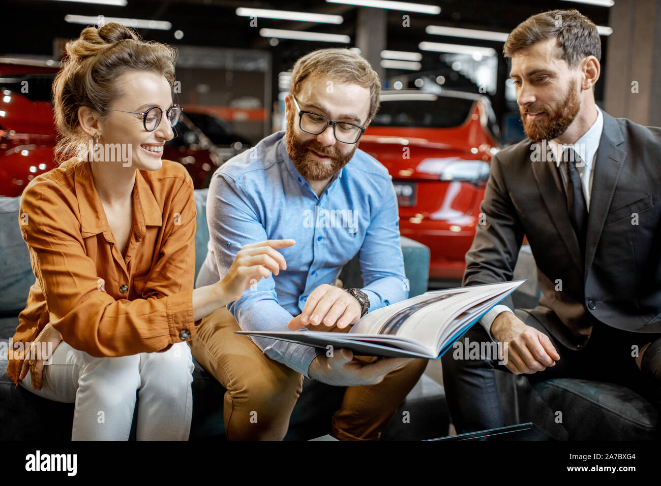 Coppia giovane la scelta di auto fine tramite un catalogo seduti con il venditore sul comodo divano presso la concessionaria auto Foto Stock