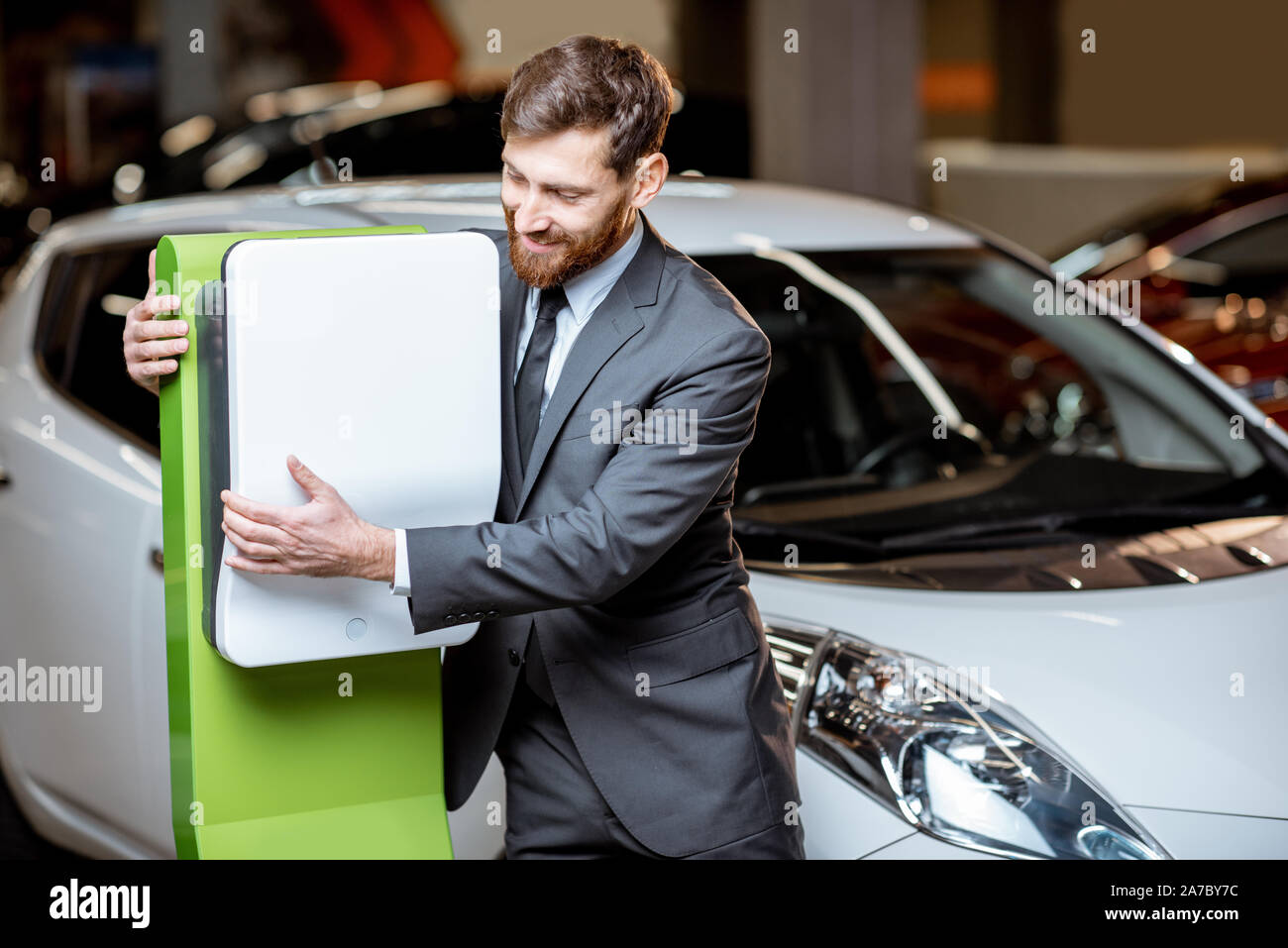 Divertente ritratto di un sales manager abbracciando auto stazione di carica nello showroom presso la concessionaria auto Foto Stock