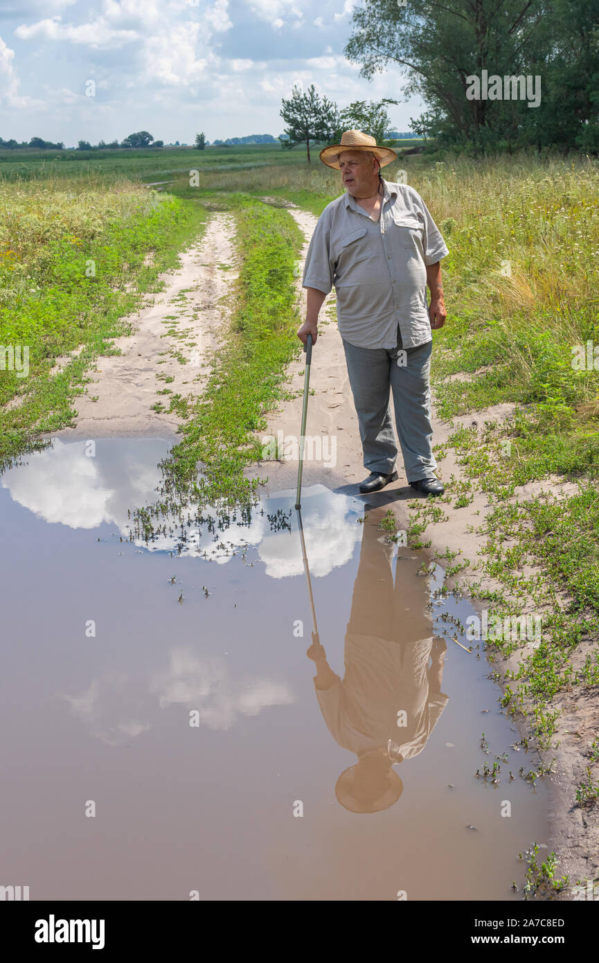 Senior uomo in hat in piedi vicino al grande pozzanghera sulla strada del paese Foto Stock