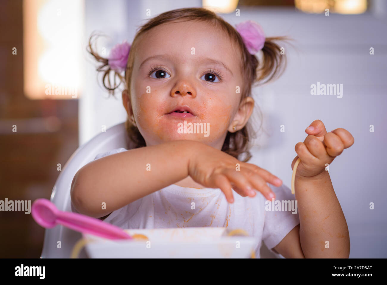 Carino piccolo funny girl di 18 mesi / 1 anno di età, baby, ragazza, seduta in una alta potenza di sedia a casa, mangia tagliatelle con cucchiaio e forchetta, all'interno di foto Foto Stock