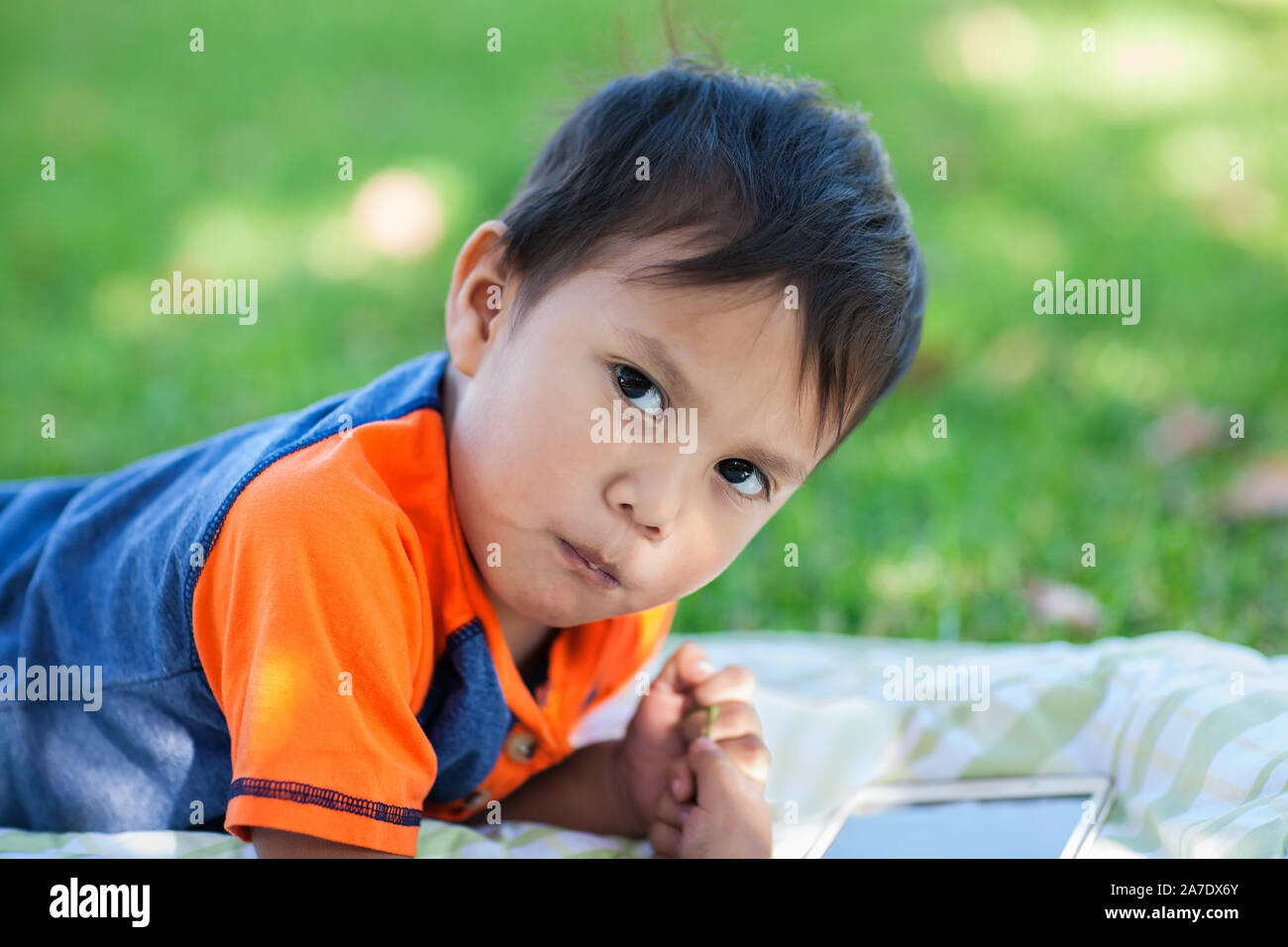 Un ragazzo latino prendendo una pausa dalla riproduzione di giochi didattici sul suo tablet, mentre recante sul prato. Foto Stock