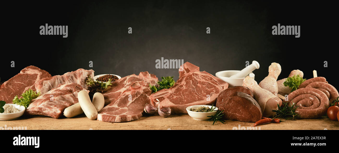 Materie assortimento di tagli di manzo, maiale, pollo e salsicce per un barbecue visualizzato con erbe aromatiche e spezie in una lunga fila su un tavolo da cucina in un panor Foto Stock