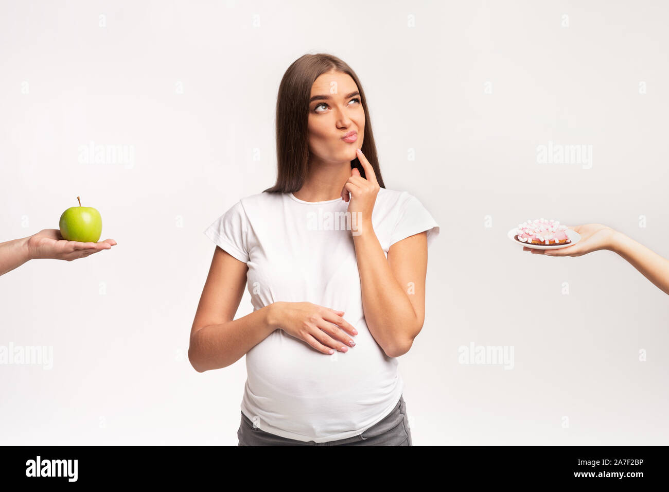 Perplesso ragazza incinta scegliendo tra Apple e la ciambella, sfondo bianco Foto Stock