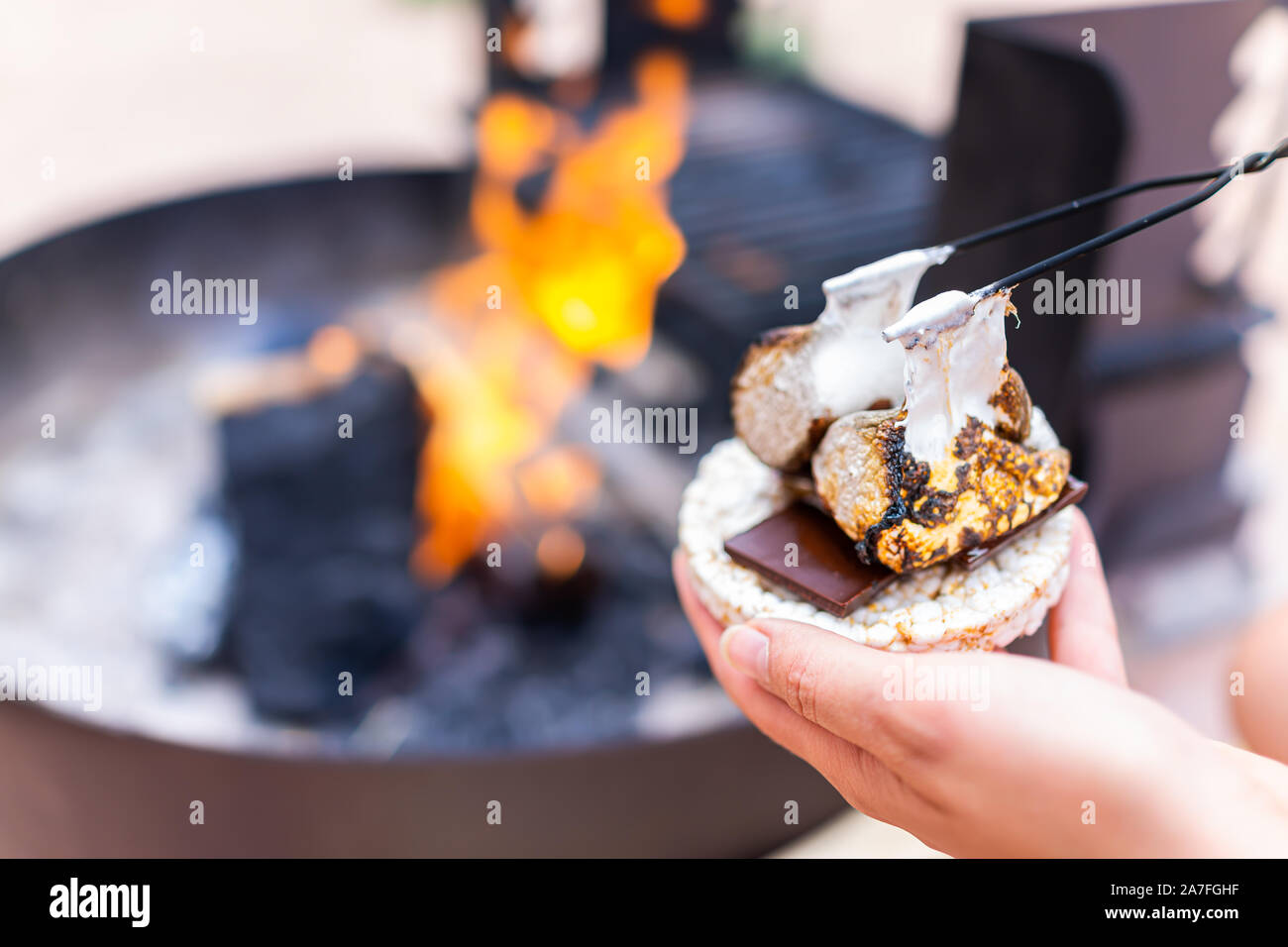 Giovane donna mano azienda gooey carbonizzati arrosto marshmallows smores con cioccolato e torta di riso cracker dal fuoco in campeggio campfire grill Foto Stock
