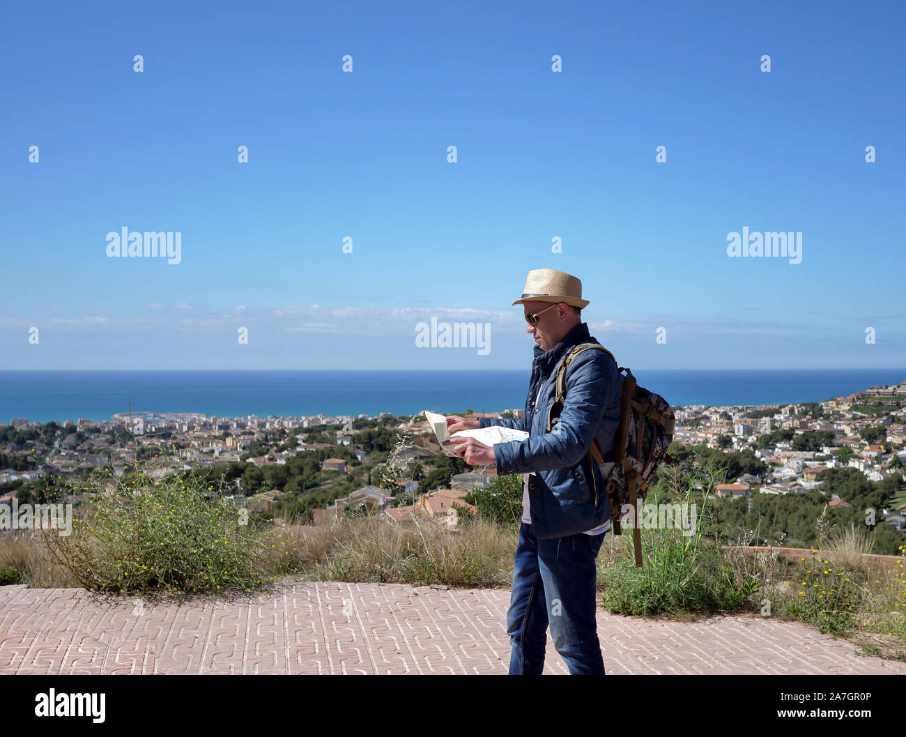 Uomo di viaggiatori su una piattaforma di osservazione sulla cima di una montagna con una mappa dell'area nelle sue mani, contro lo sfondo di una città in riva al mare Foto Stock