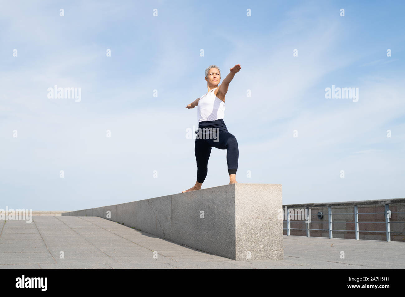 Donna di mezza età facendo due guerriero yoga pone sul muro di pietra Foto Stock