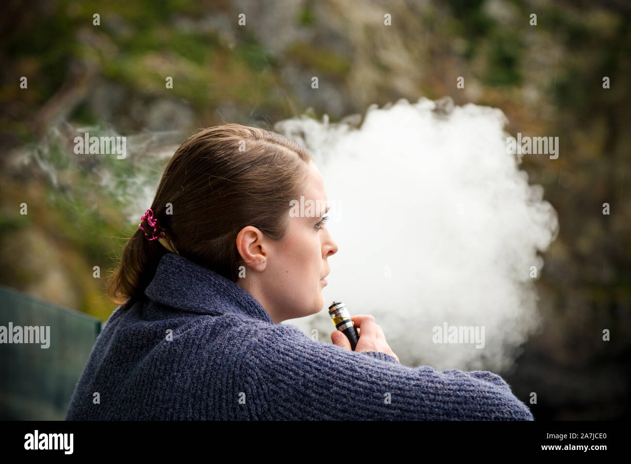 Bruna giovane donna caucasica fumare una vape ecigarette mentre in piedi su un balcone che si affaccia sull'oceano. Foto Stock