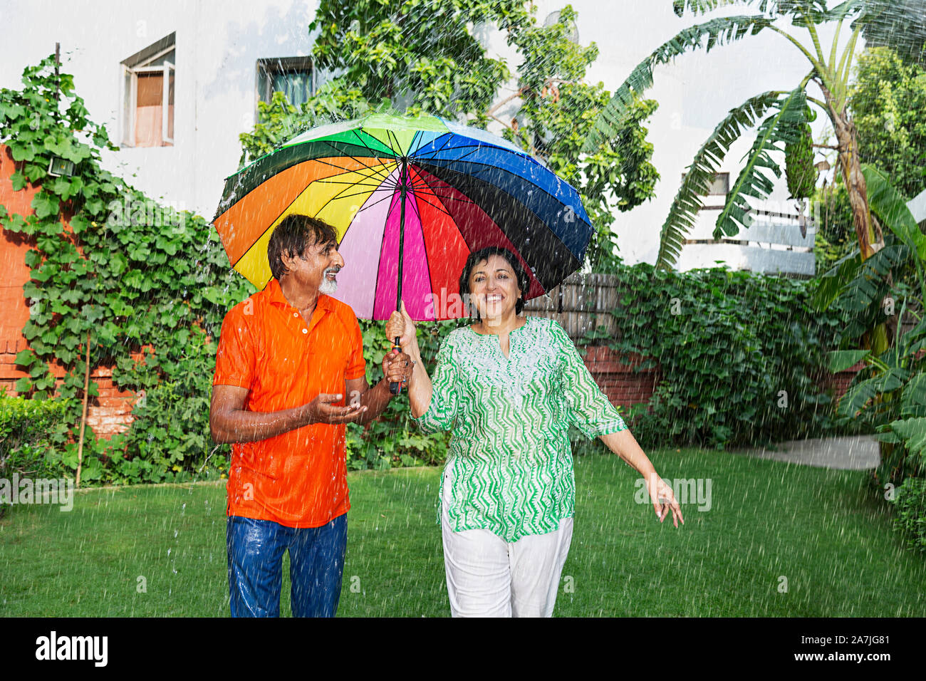 Felice Coppia di anziani con ombrellone in piedi sotto la pioggia e parlando godendo la pioggia nel giardino di casa Foto Stock