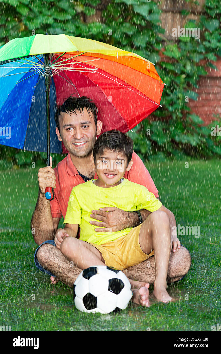 Felice famiglia indiana padre e Kid Boy sotto ombrellone seduta-su-erba con il calcio godendo della pioggia di estate in giardino Foto Stock