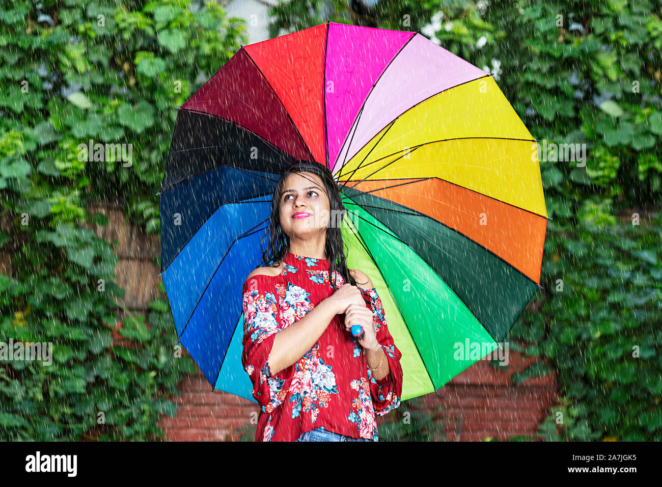 Felice femmina adulta sotto la pioggia che copre il giorno con ombrello arcobaleno in giardino esterno Foto Stock
