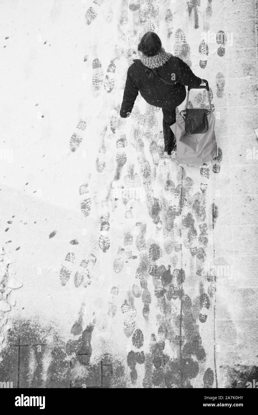 Sfocata donna camminare durante il traino di un carrello di shopping bag durante la nevicata, elevato angolo vista da dietro in bianco e nero Foto Stock
