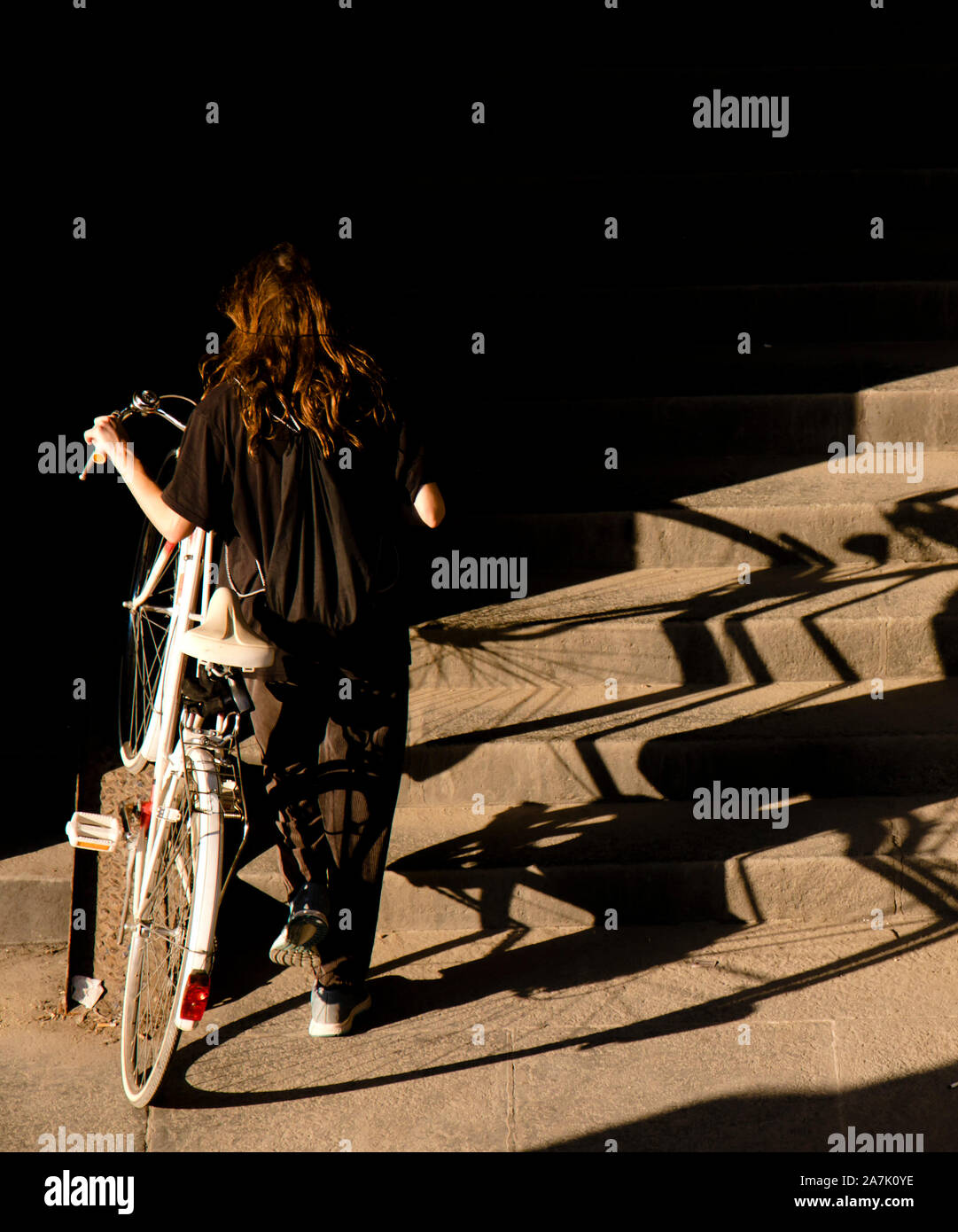 Una ragazza adolescente in nero spingendo la bici bianco fino al pubblico le scale di luce e ombre, da dietro Foto Stock
