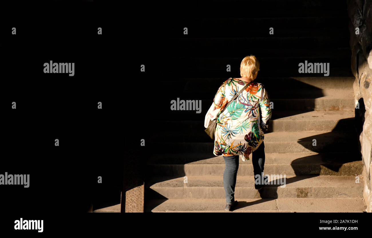 Una bionda Taglie donna a piedi fino al pubblico le scale di luce e ombre, da dietro Foto Stock