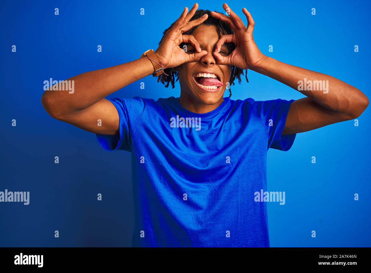 Afro-americano con dreadlocks che indossa la t-shirt in piedi isolato su sfondo blu facendo ok gesto come binocolo con la lingua fuori, gli occhi lo Foto Stock