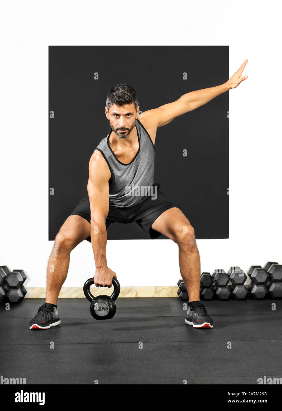 Uomo a fare gli esercizi con un peso kettlebell in una palestra sollevandolo nella posizione di squat a rafforzare i suoi muscoli glutei e gambe in salute e fitn Foto Stock