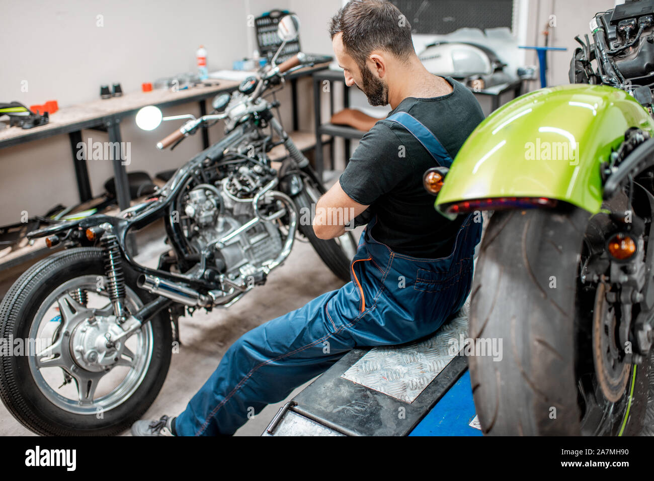 Ritratto di un bel barbuto biker in tute da lavoro seduta vicino alla moto durante la riparazione in officina Foto Stock