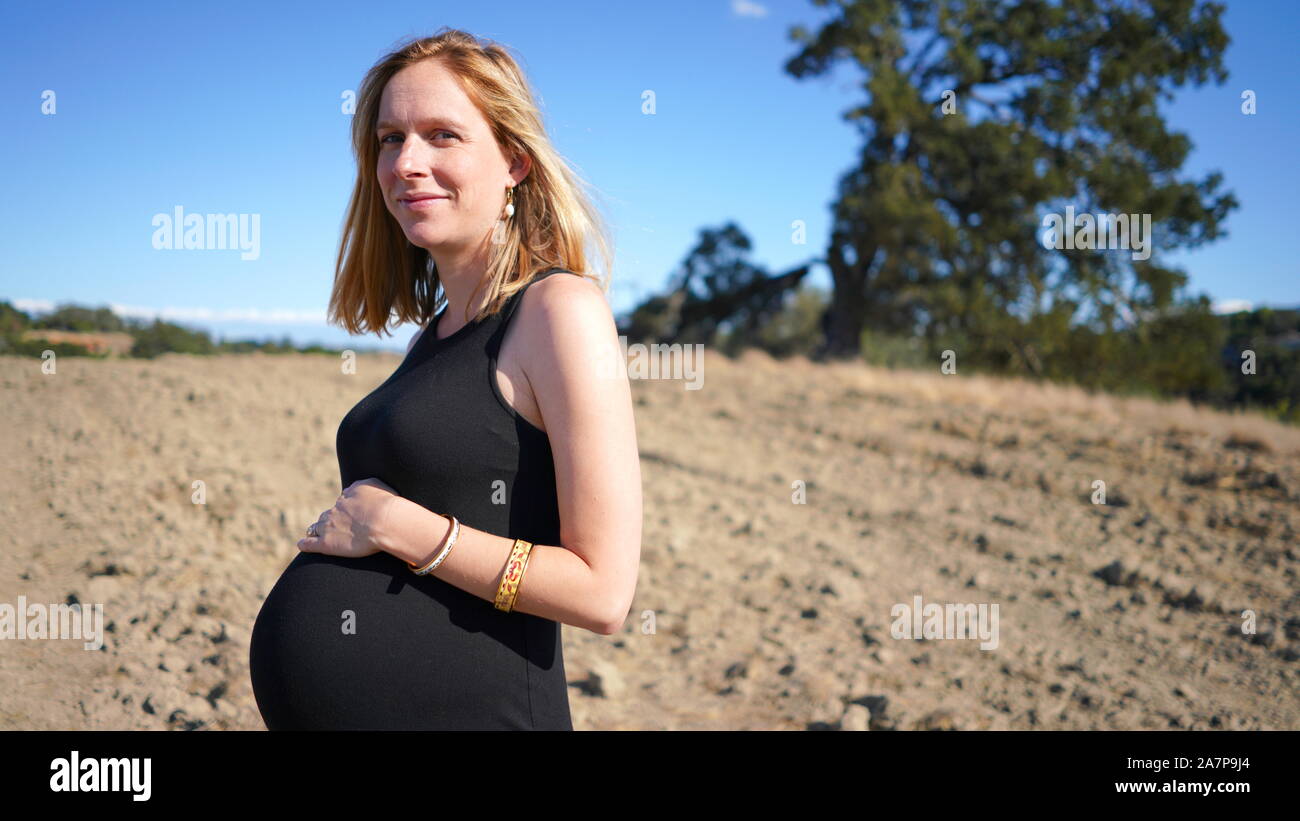 Giovane donna bionda che indossa estendibili vestito nero, ovviamente molto incinta, in piedi in primo piano del paese rurale scena di sporcizia e di massa grande albero Foto Stock