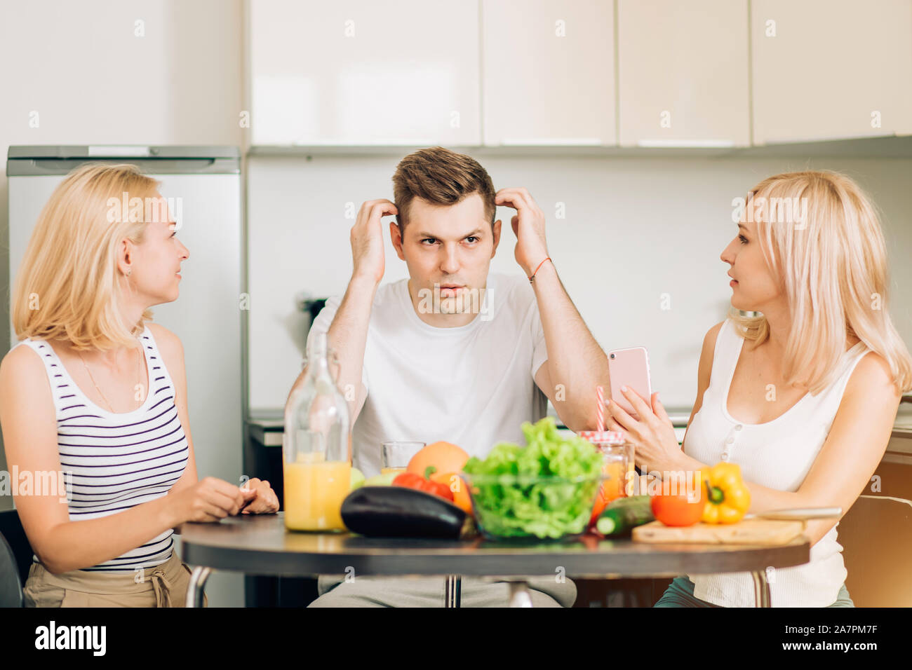 Due belle caucasica bionde ascoltando il loro amico racconta la storia interessante. Tre amici seduti a tavola in cucina, potabile e frutta veget Foto Stock