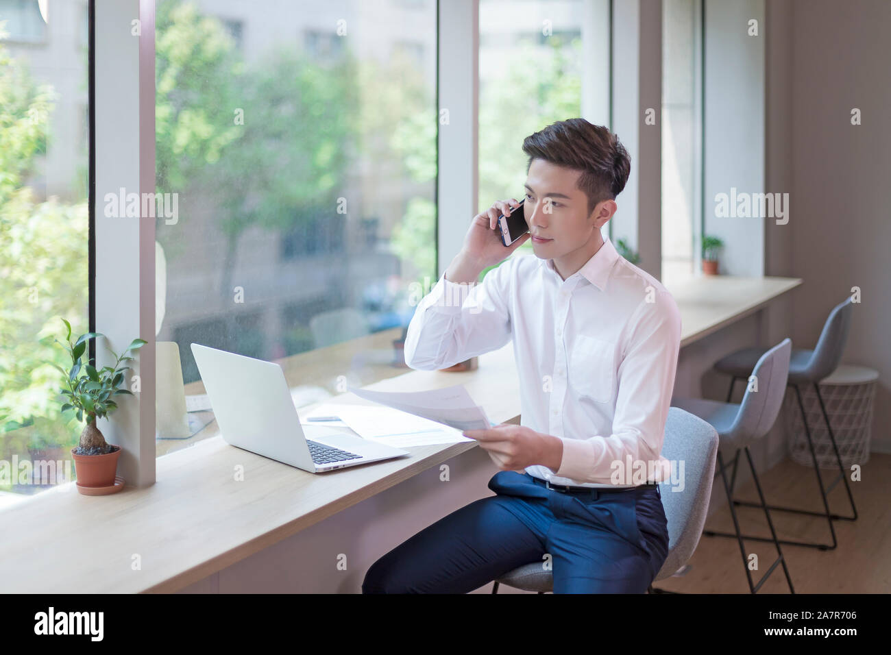Giovane imprenditore con corti capelli neri e un bianco button down shirt parlando al telefono a una scrivania in un ufficio mentre si tiene un documento Foto Stock