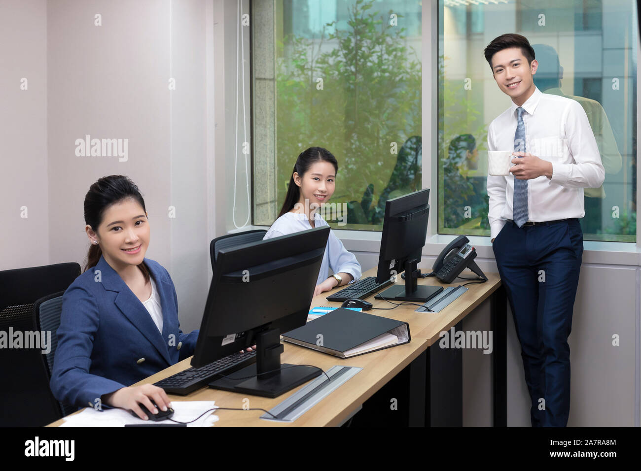 Ritratto di tre giovani uomini e donne sorridenti che guardano la macchina fotografica in un ufficio con due di loro seduti ai loro computer Foto Stock