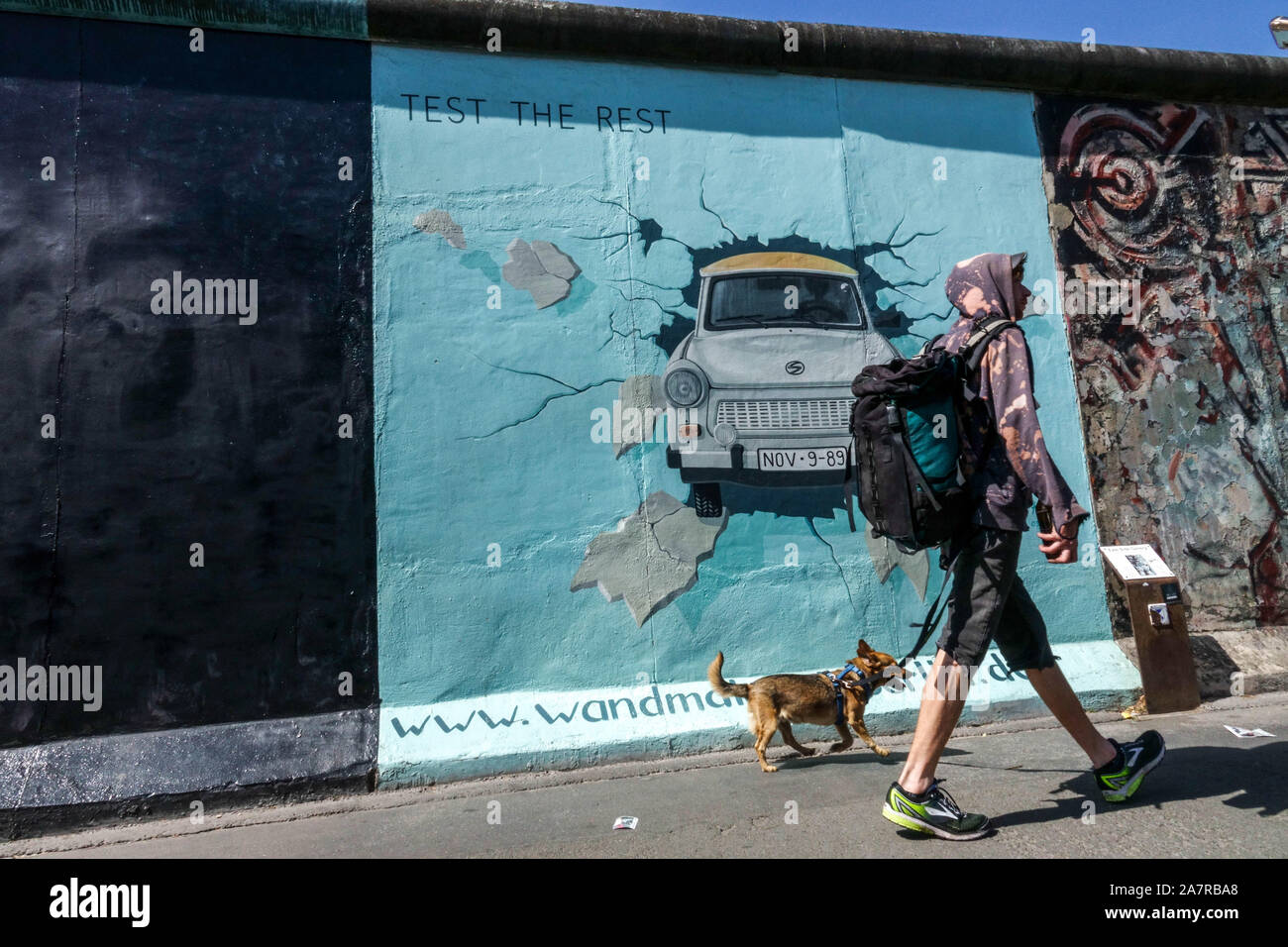 Berlino muro graffiti East Side Gallery Germania giovane uomo a piedi Il cane che passa intorno Trabant auto Friedrichshain città strada Berlino Persone in città Foto Stock