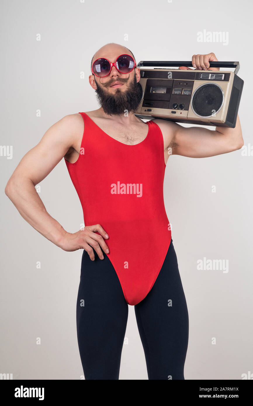 Ritratto di un divertente uomo barbuto in un body rosso e nero leggings tenendo un retrò registratore a nastro su sfondo bianco Foto Stock
