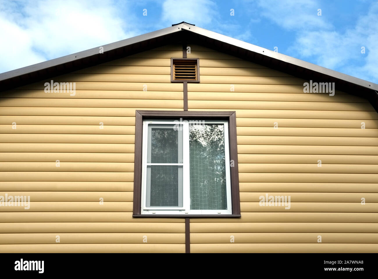Piano superiore con finestra in plastica in country house parete ricoperta con schierandosi giallo e marrone e tetto metallico sul cielo blu con nuvole sulla giornata soleggiata vista frontale Foto Stock