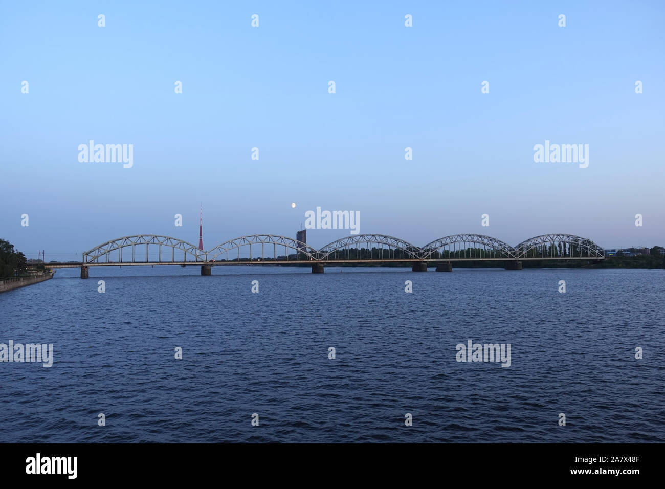 Metallo lungo la ferrovia ponte ad arco attraverso il fiume Daugava a Riga contro stand alone edificio, la torre della TV e luminosa luna vista in avanti sul Cielo di tramonto Foto Stock