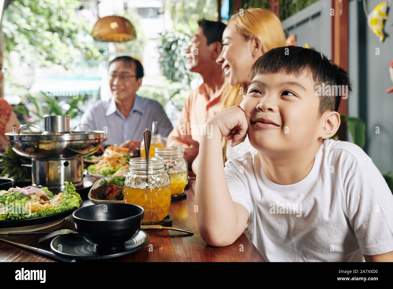 Adorabili poco ragazzo vietnamita sorridente, guardando verso l'alto e sognare quando si è seduti al tavolo per la cena durante la festa di famiglia Foto Stock