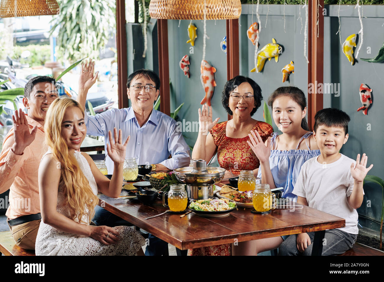 Felice grande famiglia vietnamita seduti al tavolo per la cena con il tradizionale cibo asiatico, salutando con le mani e guardando la fotocamera Foto Stock