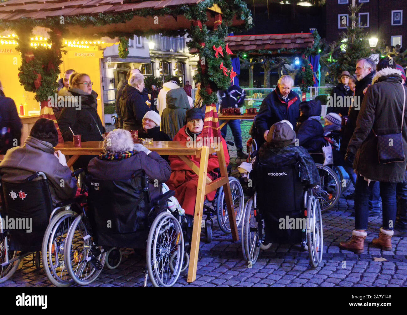 Le donne anziane con mobilità limitata in carrozzina tramite braccio muscoloso forza bere vino brulé circondato da parenti. Natale a piedi in città Foto Stock