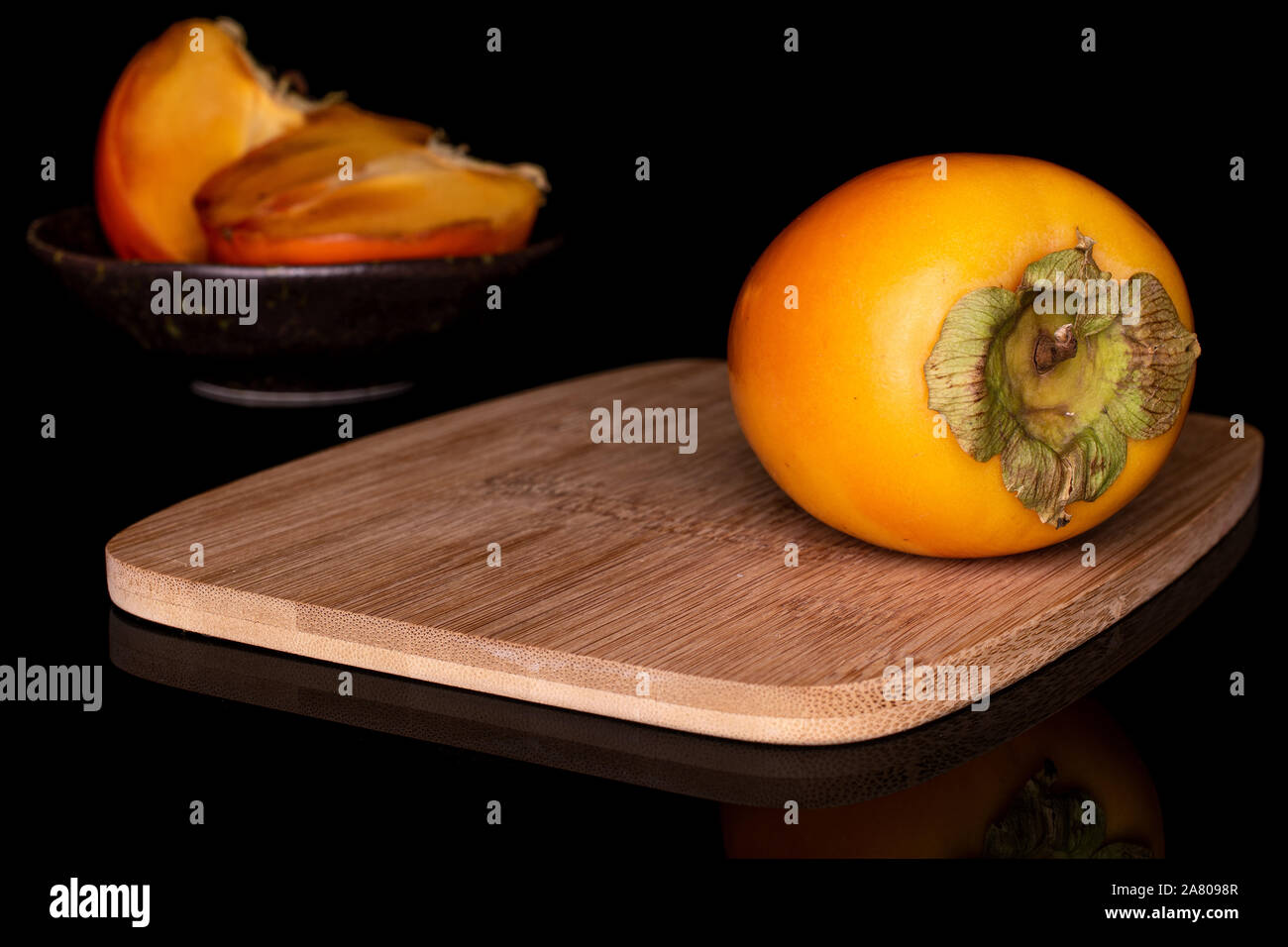 Gruppo di una intera due metà di arancio dolce persimmon nel recipiente smaltato sul bambù tagliere isolato sul vetro nero Foto Stock