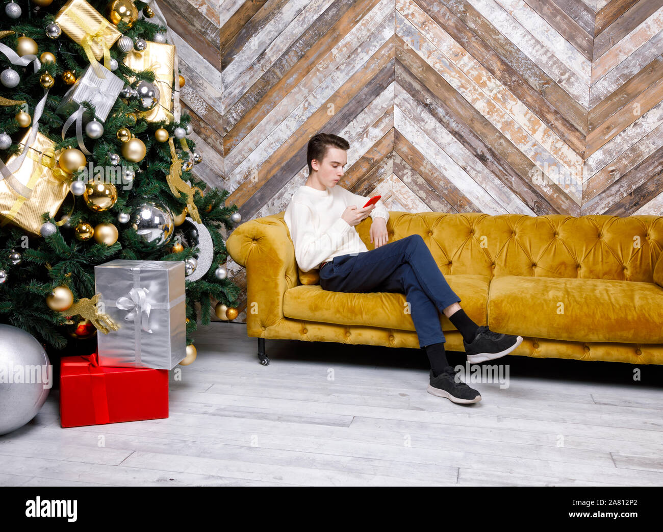 Adolescente ragazzo seduto da solo su un divano accanto ad albero di natale con doni si è concentrato solo sul suo smartphone messaging o il controllo di social media - Smombie holi Foto Stock