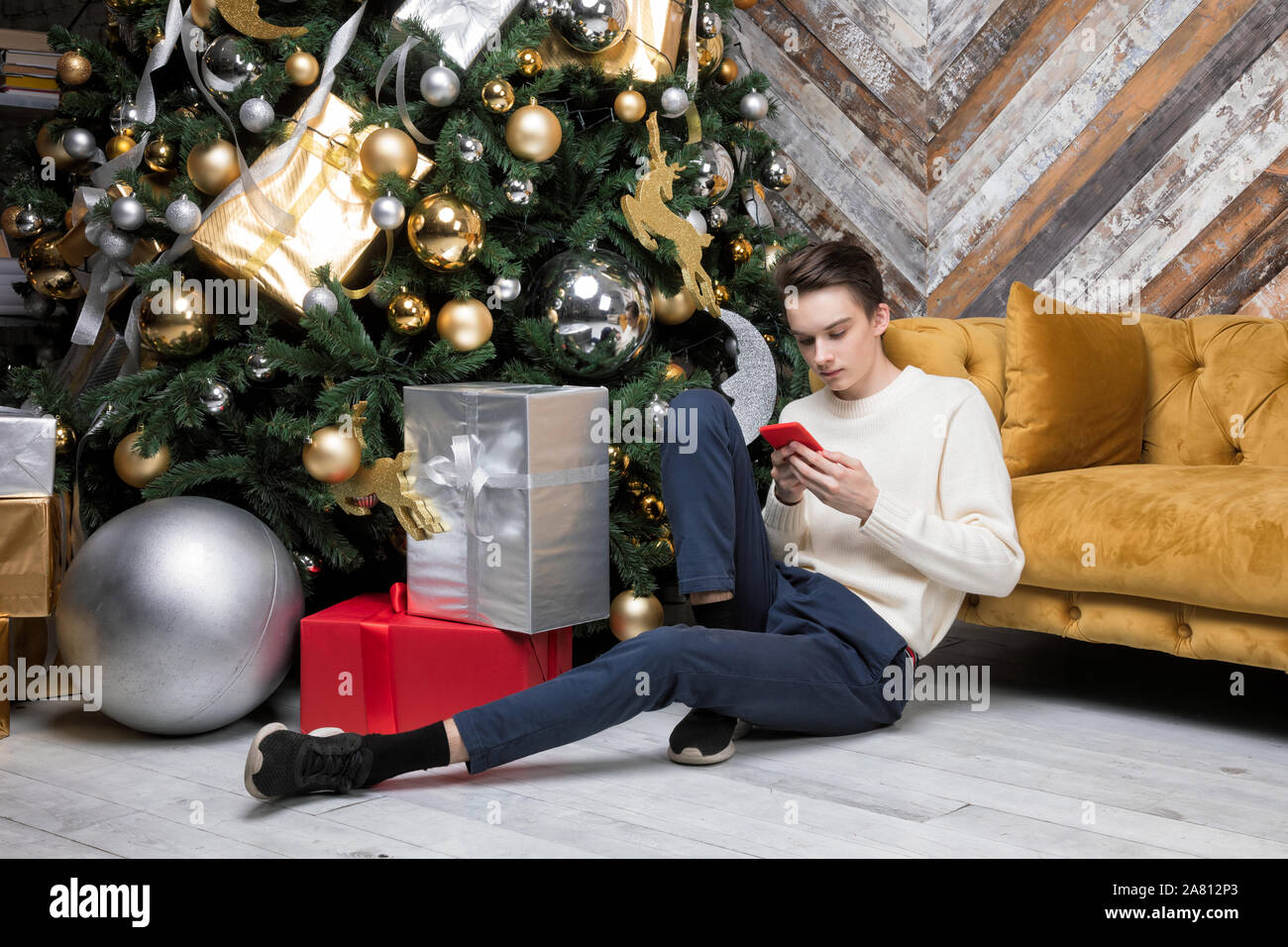 Adolescente ragazzo seduto da solo su un pavimento da albero di natale con doni si è concentrato solo sul suo smartphone messaging o il controllo di social media - Smombie holiday Foto Stock