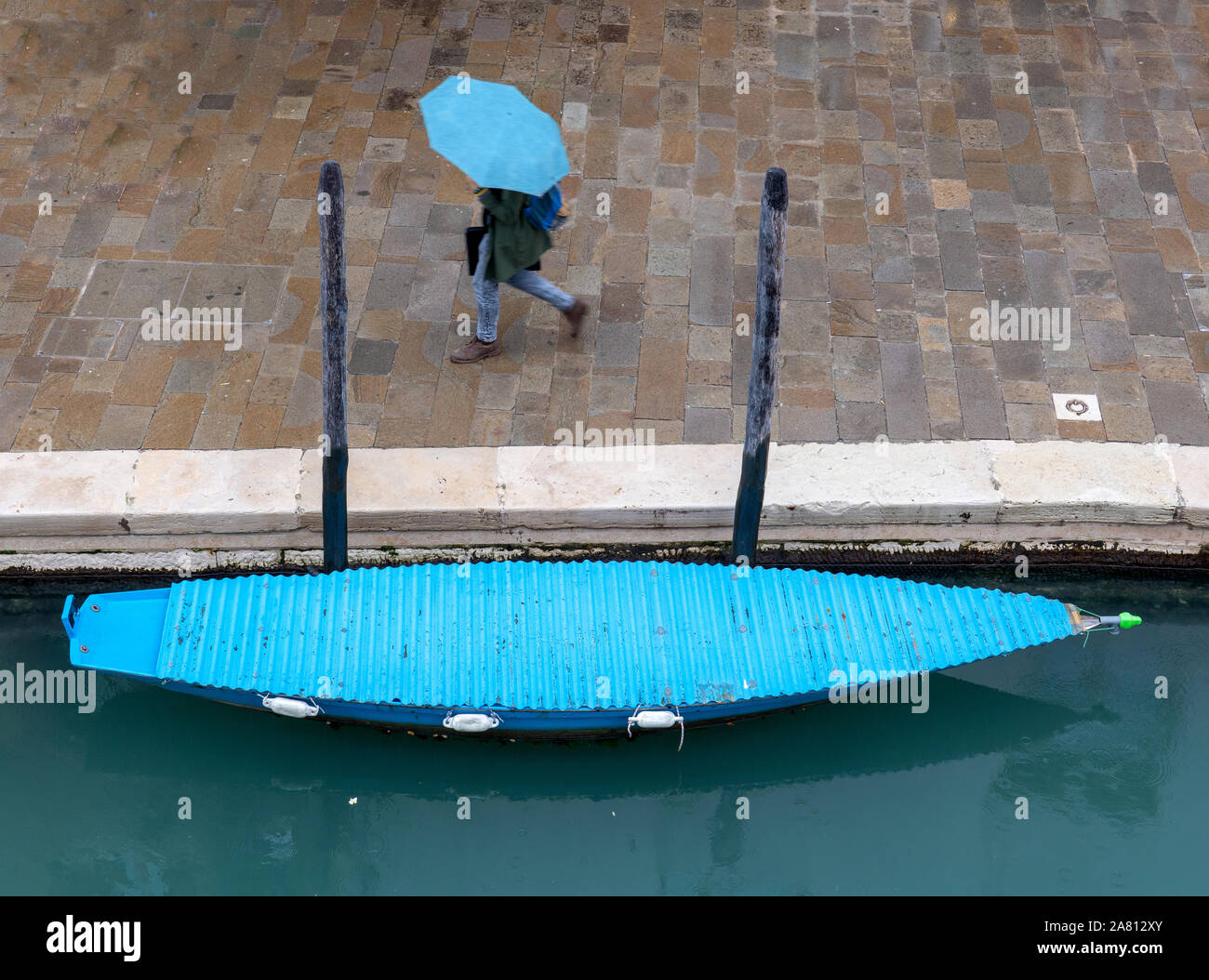 Un uomo con un ombrello blu oltrepassando una barca con un coperchio blu da un canale di Venezia Italia Foto Stock